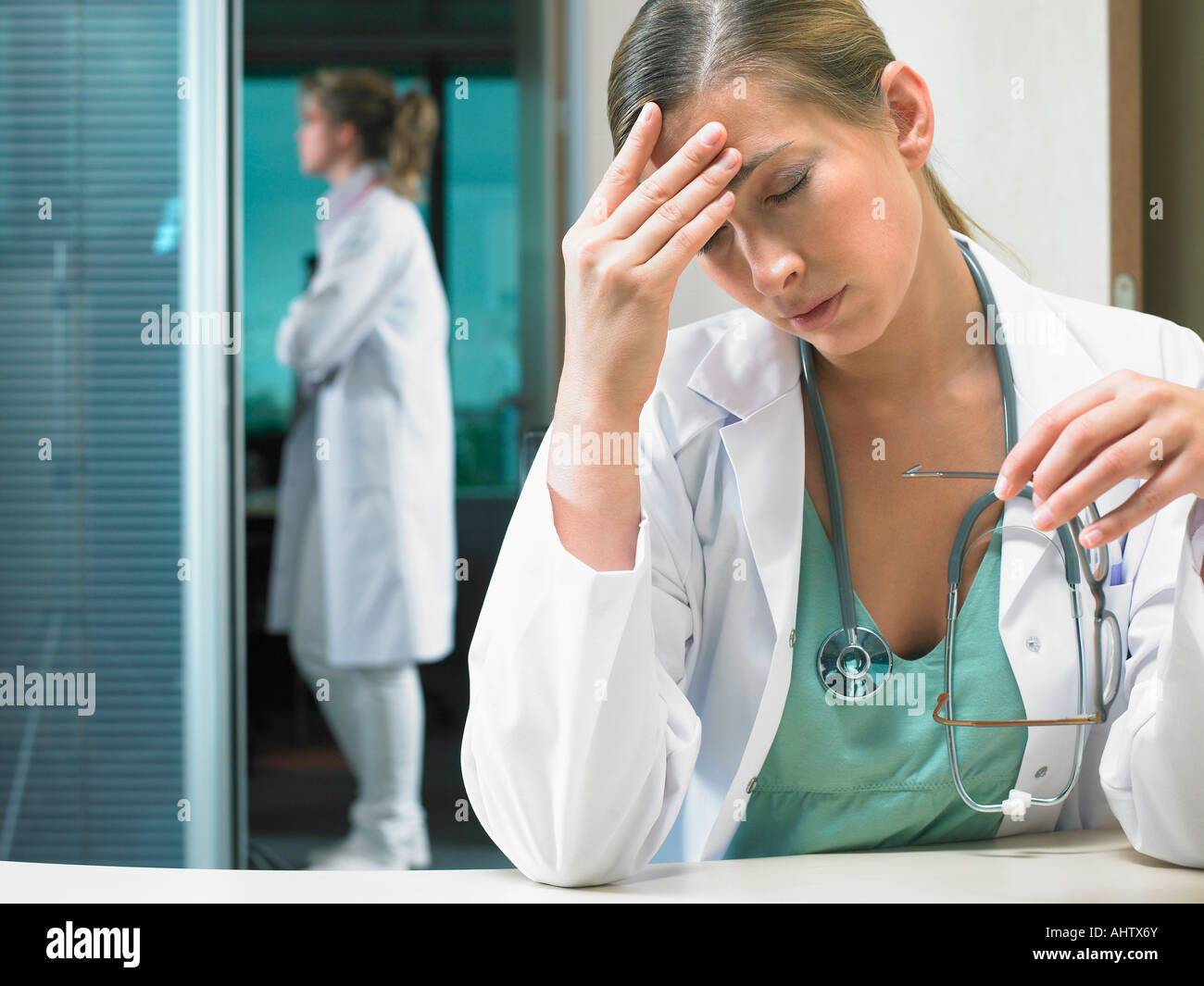 Müde Arzt sitzt an ihrem Schreibtisch mit Ärztin im Hintergrund stehen. Stockfoto