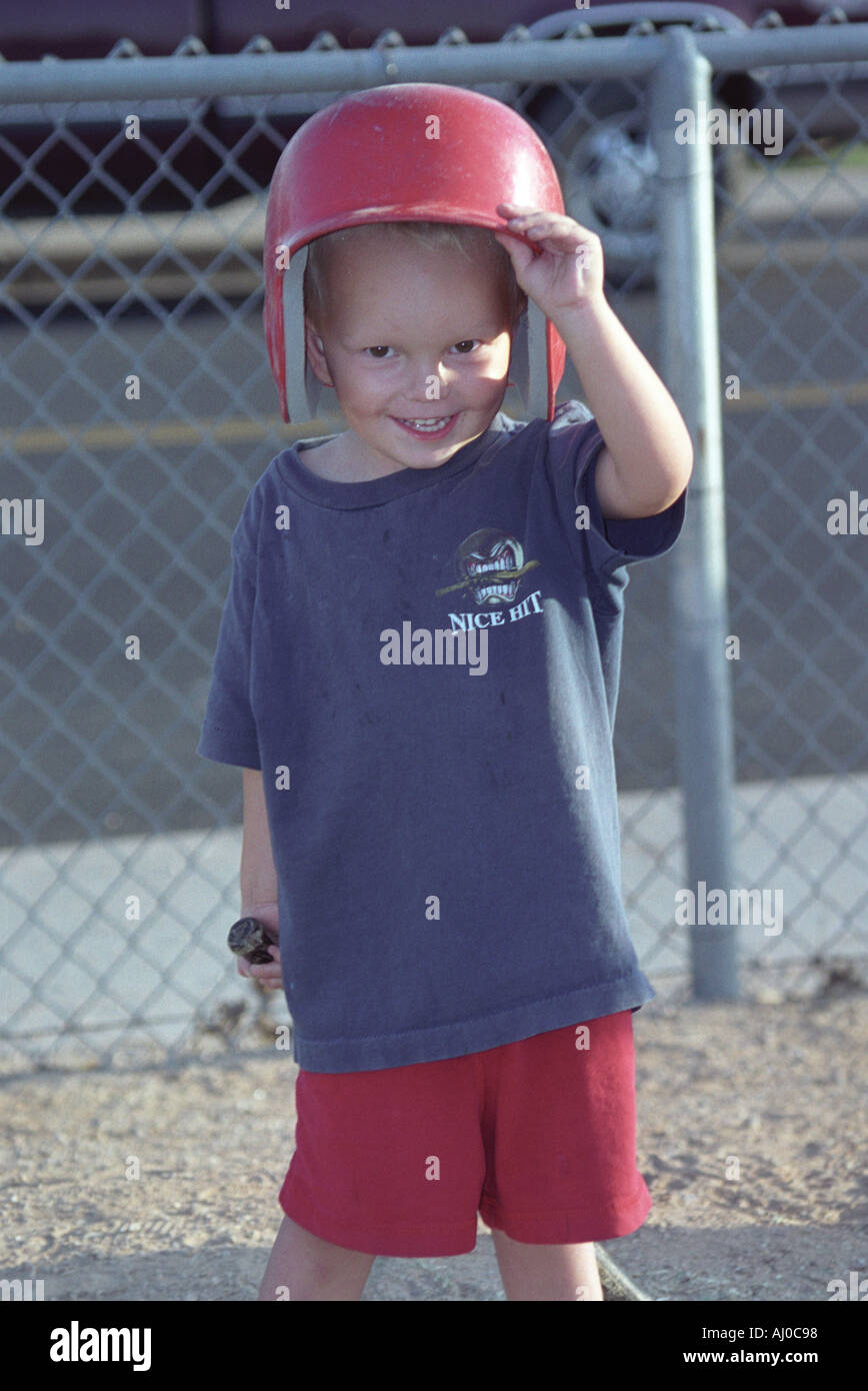 Ein Lächeln auf den Lippen kleiner Junge hebt einem Wimper Helm mehrere Größen zu groß Weg von seinen Augen als er zum Jahresende einen Baseballschläger hält Stockfoto