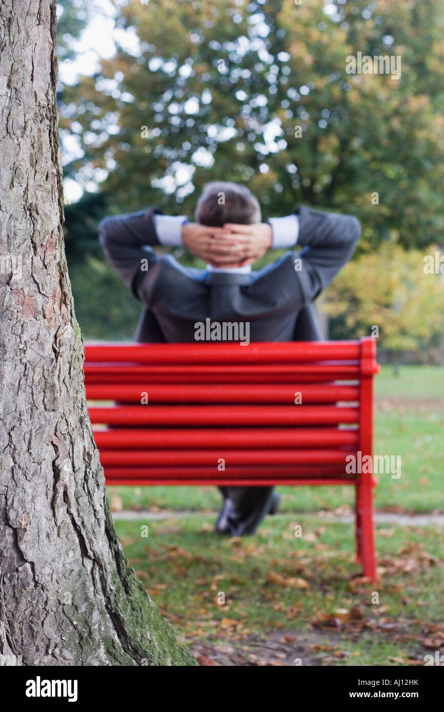 Entspannen Sie sich auf einer roten Parkbank Fokus auf dem Baum im Vordergrund Geschäftsmann Stockfoto