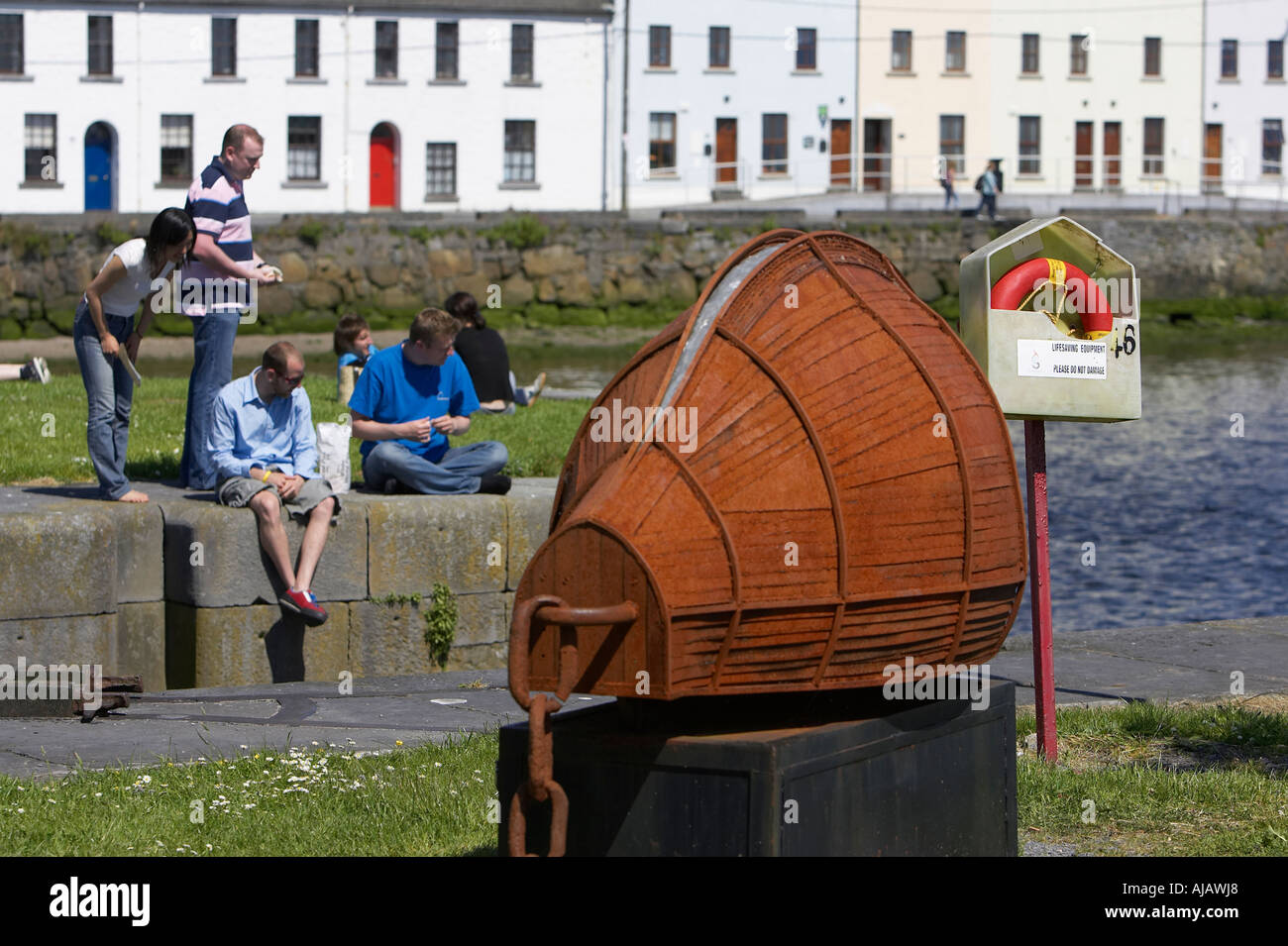 Menschen sitzen auf Pier Wand mit Rettungsring und rostigen Boot geformte Skulptur claddagh Stockfoto