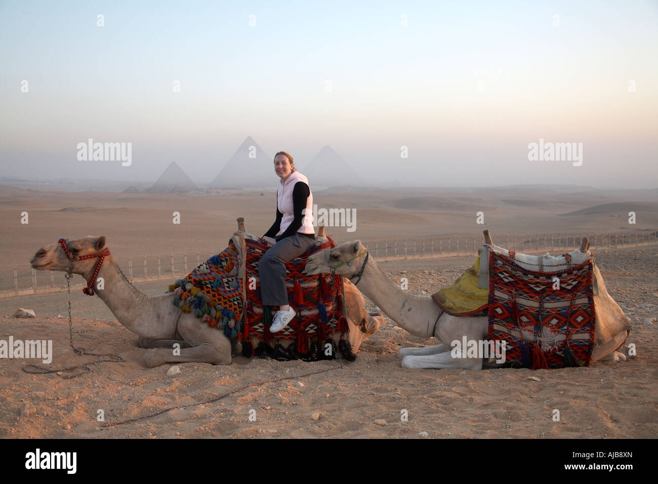 Lächelnde Frau Touristen auf einem Kamel in der Steinwüste im nebligen dunstigen Sonnenlicht am frühen Morgen mit Pyramiden in der Ferne Gizeh-Kairo Stockfoto