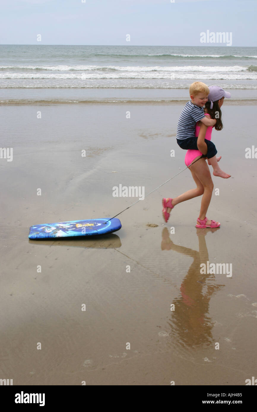 Mädchen 8-10 Jahre geben wählen Sie eine zurück zur kleinen Jungen bei einem Spaziergang entlang der Küste ziehen Surfbrett im Pembokeshire wales Stockfoto