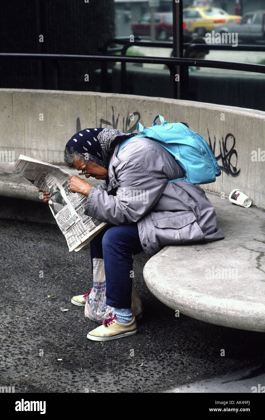 Eine ältere Frau mit einem Rucksack liest eine Zeitung auf einer Stadt-Bank Stockfoto