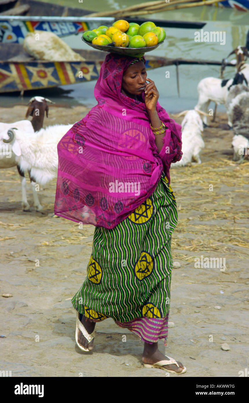 Eine Frau in traditioneller Kleidung gleicht Frucht auf dem Kopf, wie geht sie an den Ufern des Flusses Niger in Mopti Mali Stockfoto