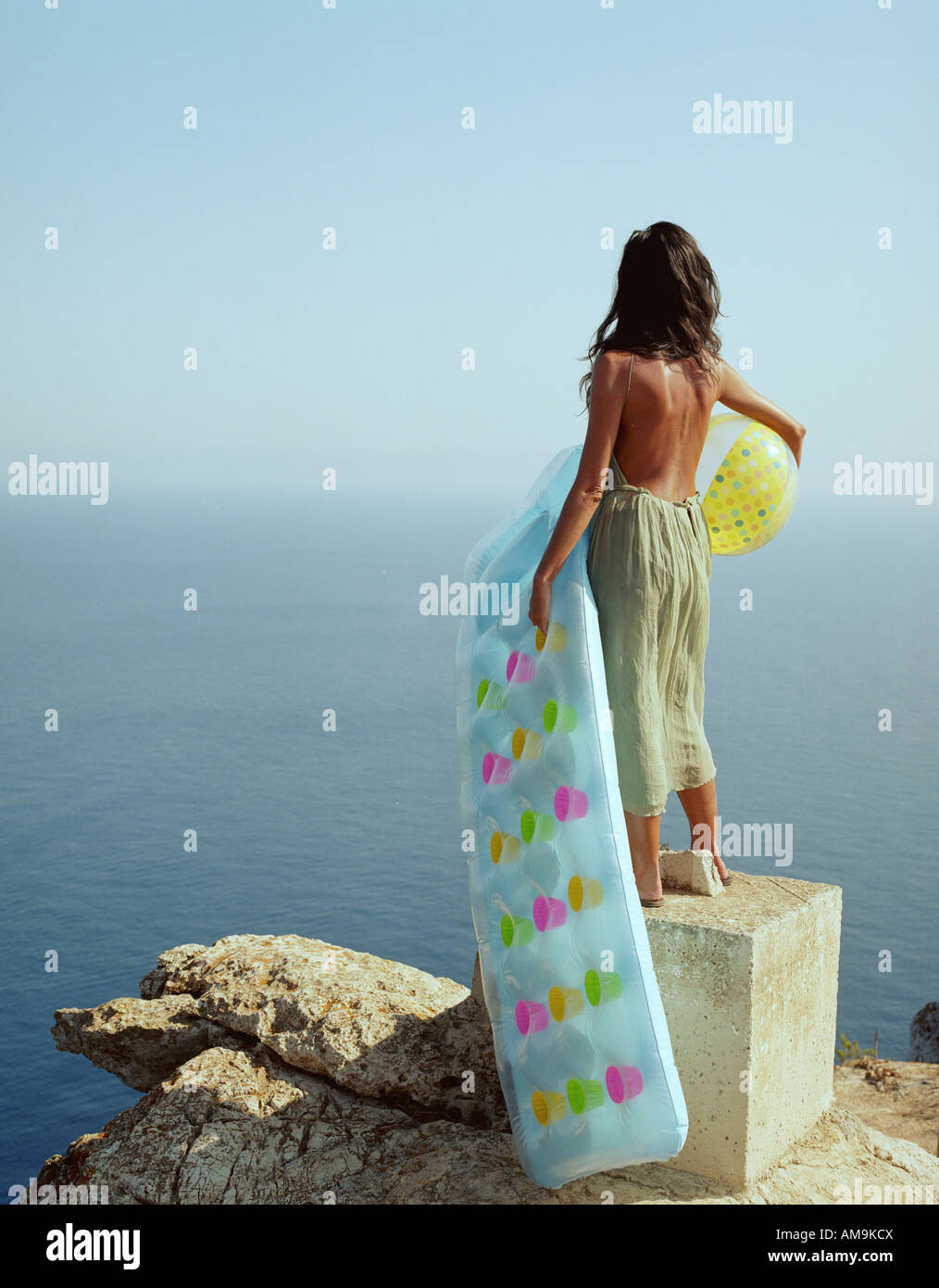 Frau auf Felsen mit Blick auf das Meer hält ein Beach-Ball und ein Lilo steht. Stockfoto