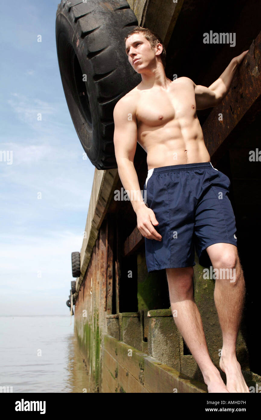 Junger Mann in Boardshorts mit gut definierten Muskeln stehen auf dem Steg am Strand an einem Sommertag. Stockfoto