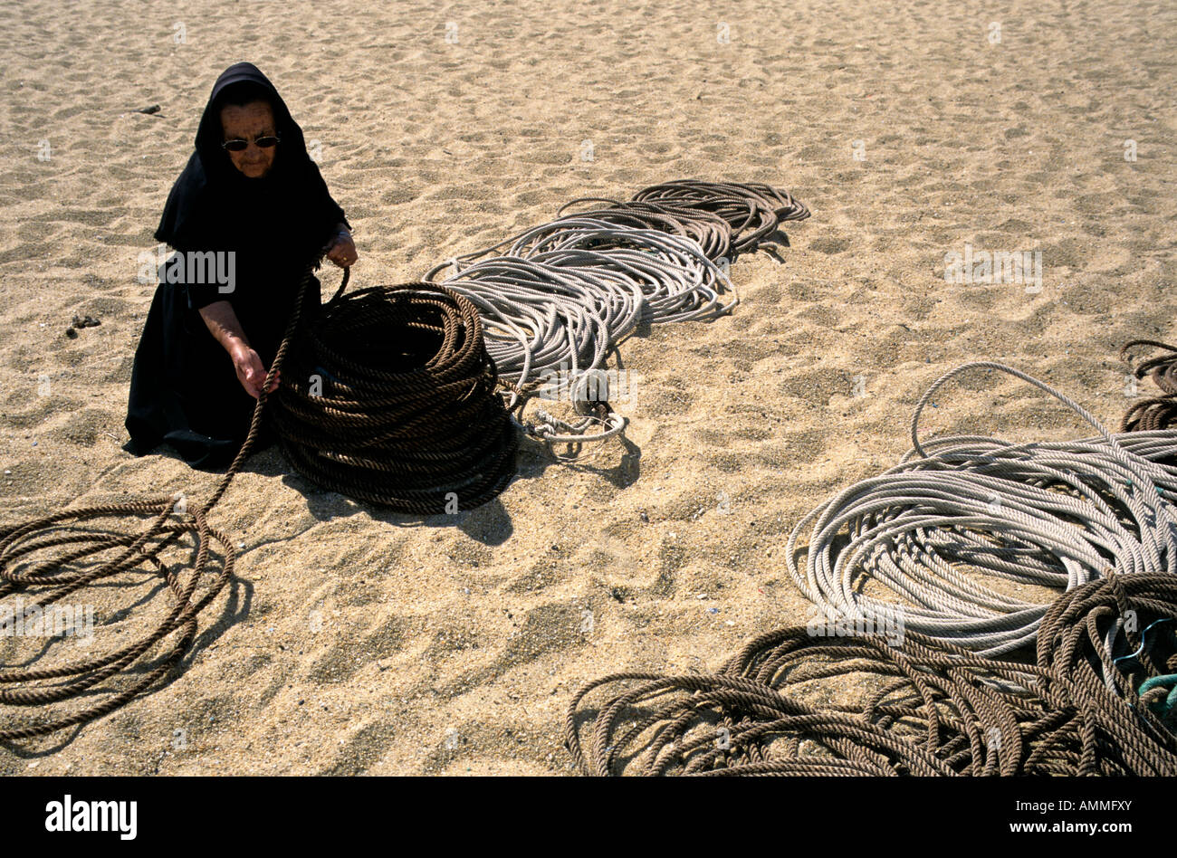 Eine alte portugiesische Frau, Head-to-Toe in schwarz gekleidet, Wicklung Seil in ordentlich Schleifen auf einem Strand von Nazaré Stockfoto