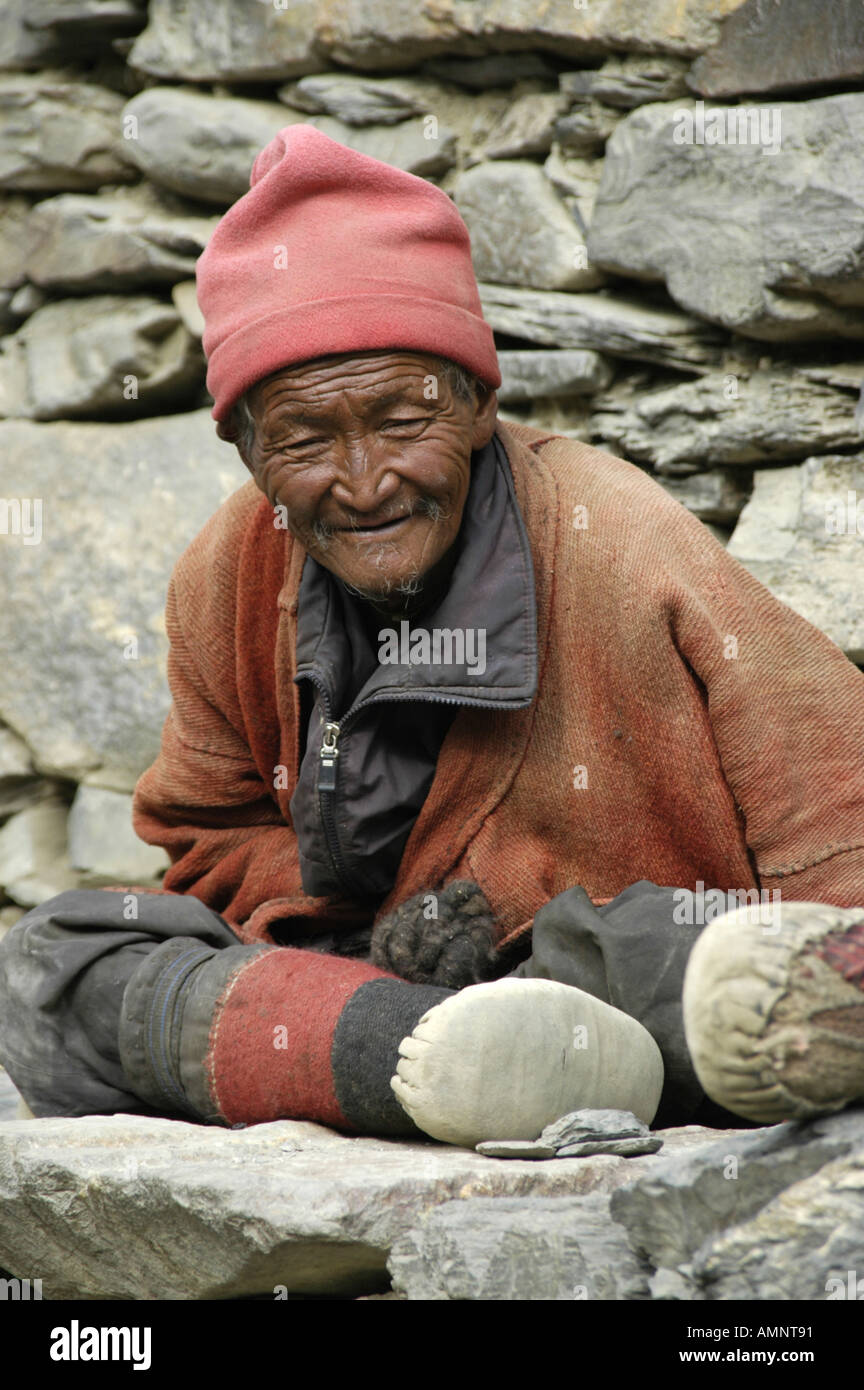 Alter Mann trägt Fleece Mütze und Umhang und Yak Leder Stiefel gegen eine Wand aus Steinen Phu Nar-Phu Annapurna Region Nepal Stockfoto