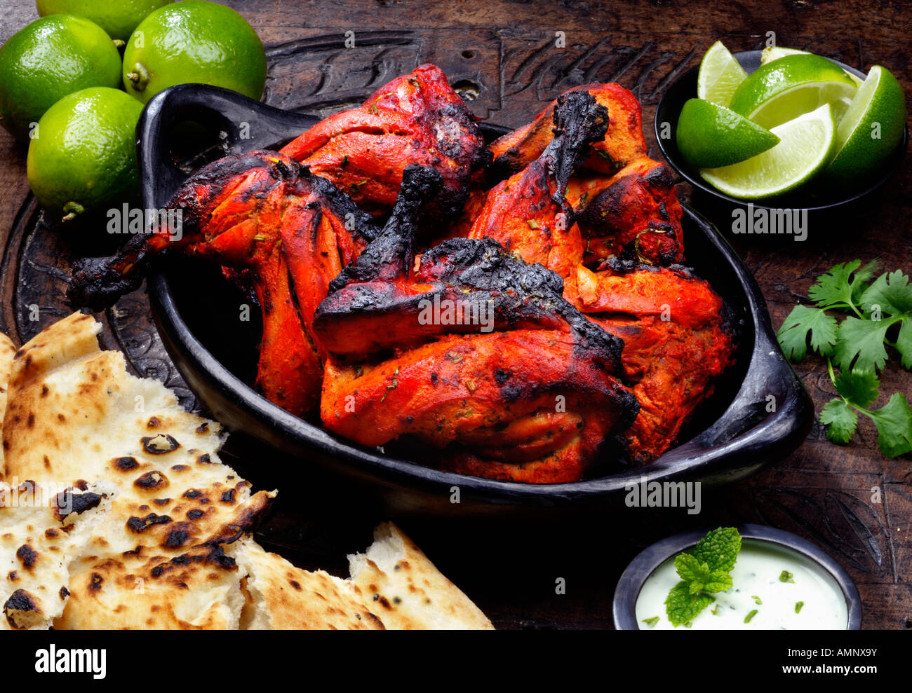 Tandoori Huhn Stücke in eine Schüssel mit Limetten, Koriander, Naan-Brot und Joghurt und Minze Dip sitzen auf einem hölzernen Hintergrund. Stockfoto