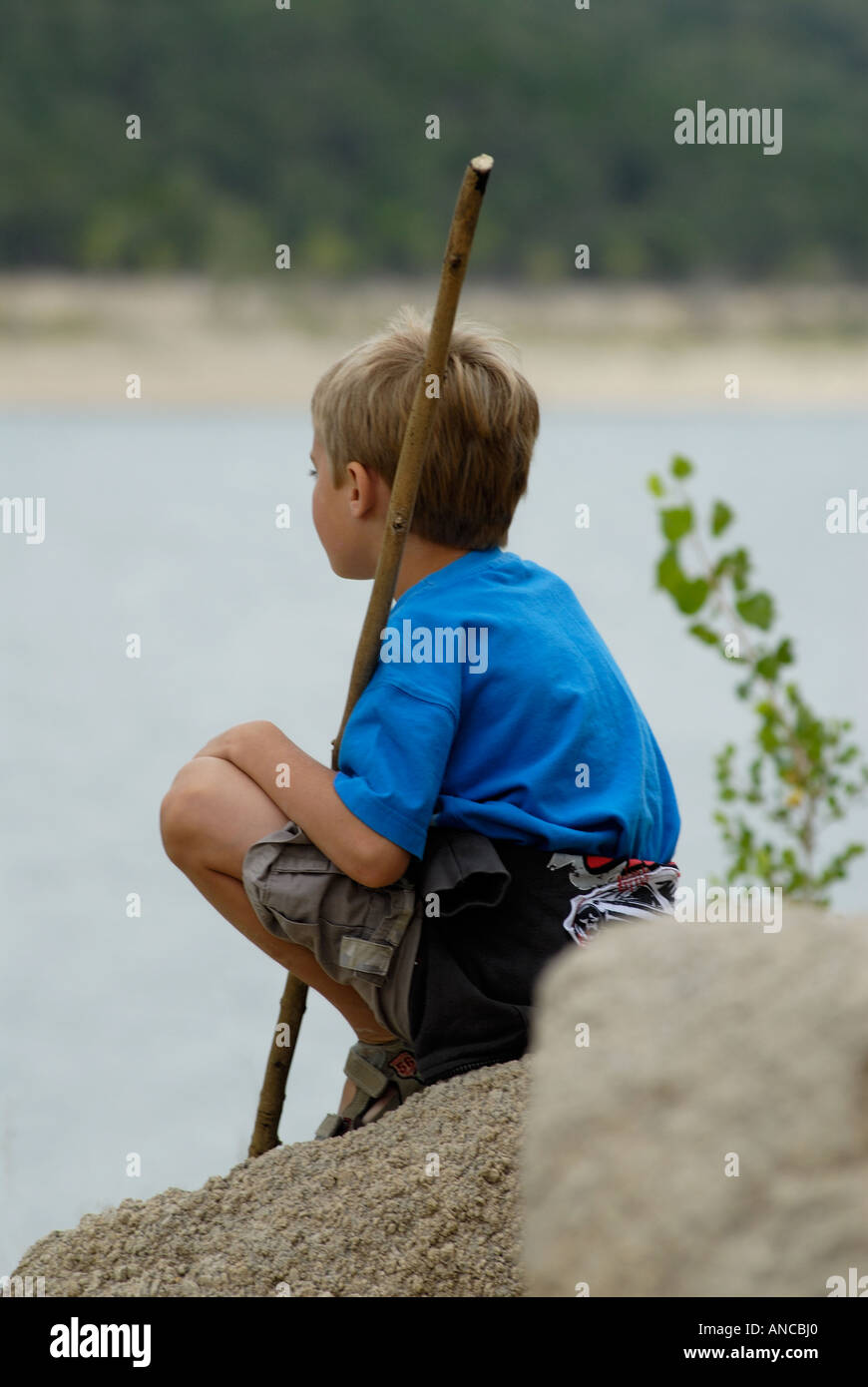 Kleiner Junge sitzt auf einem Felsen mit einem Stock, Blick auf einen See Stockfoto
