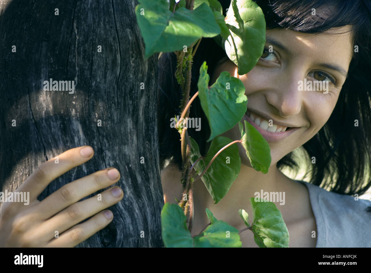 Junge Frau hinter Baumstamm, lächelnd in die Kamera, Porträt Stockfoto