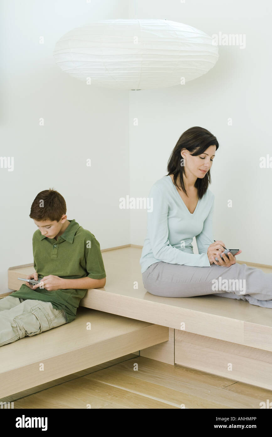 Frau und Sohn sitzen nahe beieinander, Frau mit PDA, junge spielt mit Handheld-Videospiel Stockfoto
