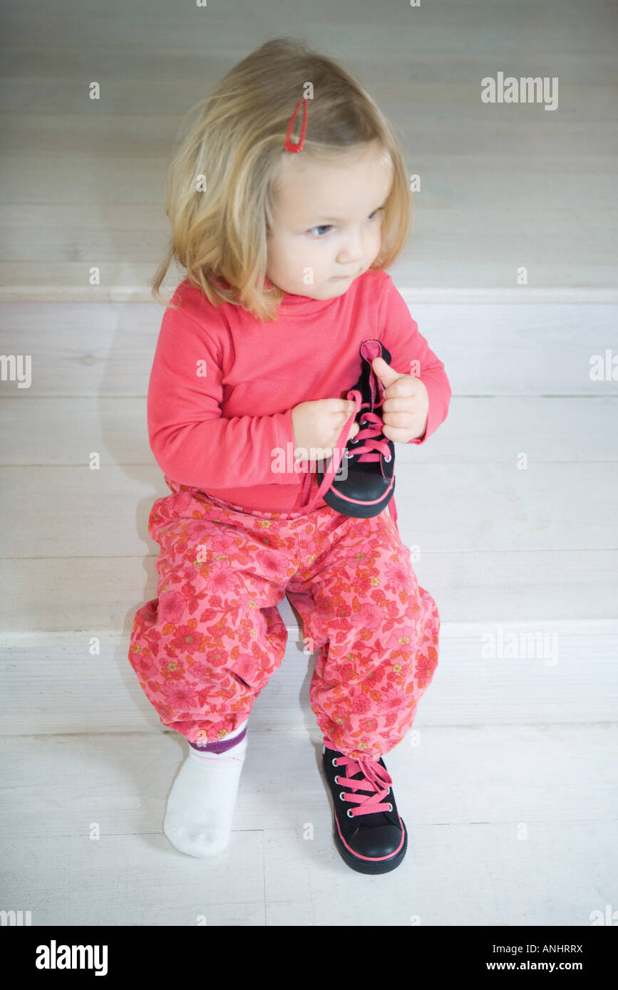 Blonde Kleinkind Mädchen sitzen auf Schritte, einen Schuh tragen, halten andere Schuh in der hand Stockfoto