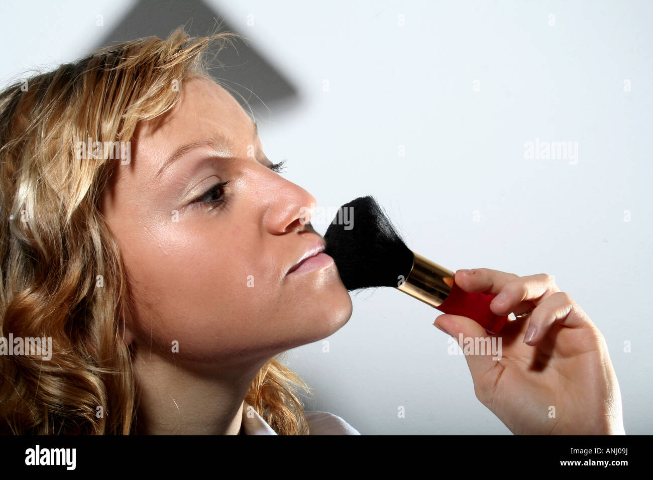 Nahaufnahme einer Frau, die Make-up mit einem weichen Pinsel aufträgt, mit Fokus auf ihre Wange, mit einem sauberen weißen Hintergrund und sanfter Beleuchtung. Stockfoto