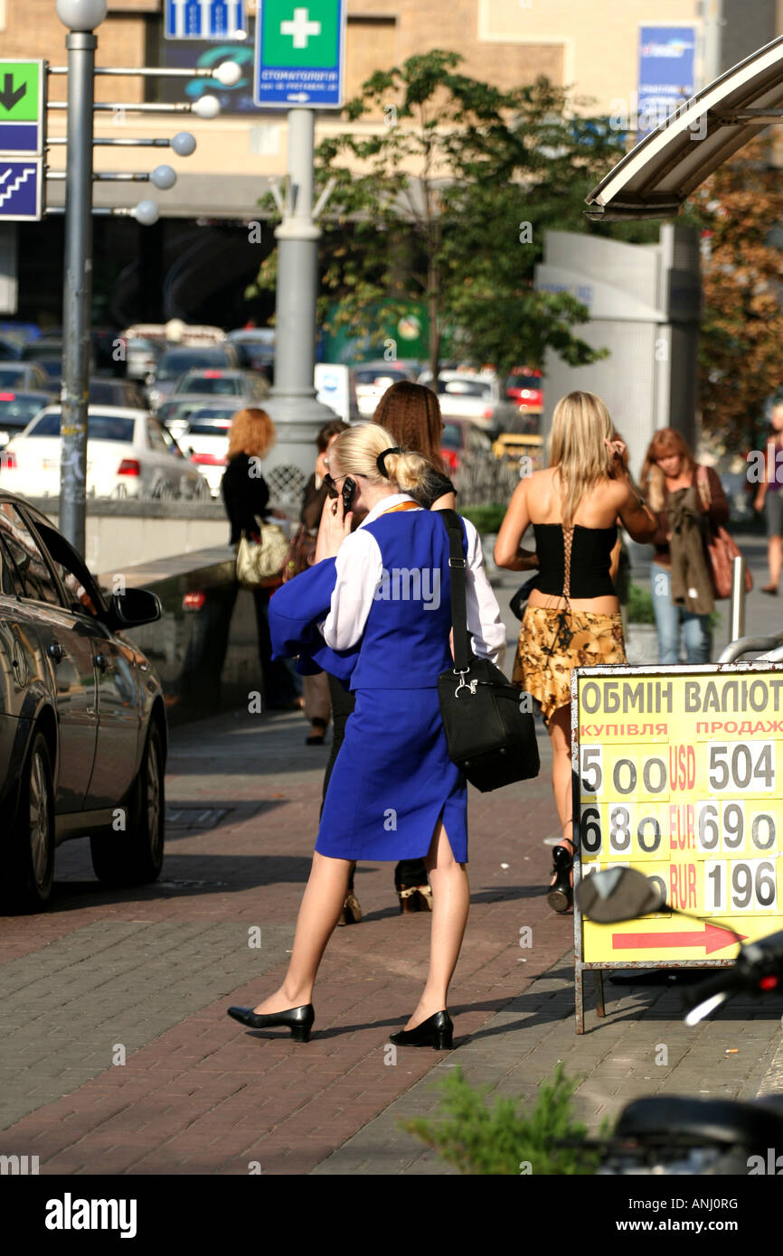 Frauen, die auf einer belebten Straße in Kiew laufen, darunter eine in blauer Uniform, mit Autos, Schildern und Wechselkursen im Hintergrund. Stockfoto