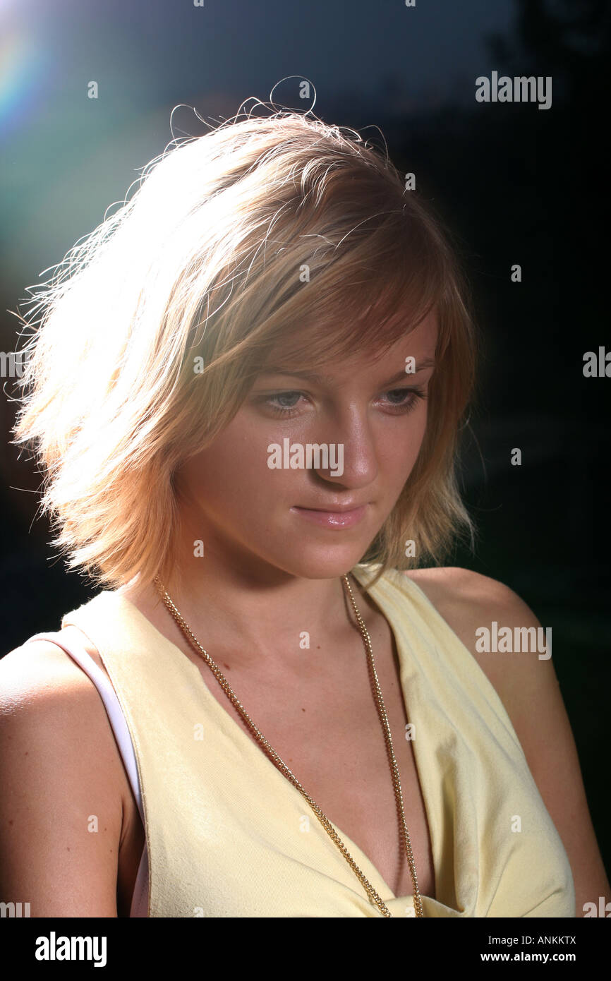 Blonde Frau mit gelbem Oberteil und gelber Halskette, nachdenklich nach unten blickend, beleuchtet durch sanftes Licht, was eine ruhige Stimmung schafft. Stockfoto