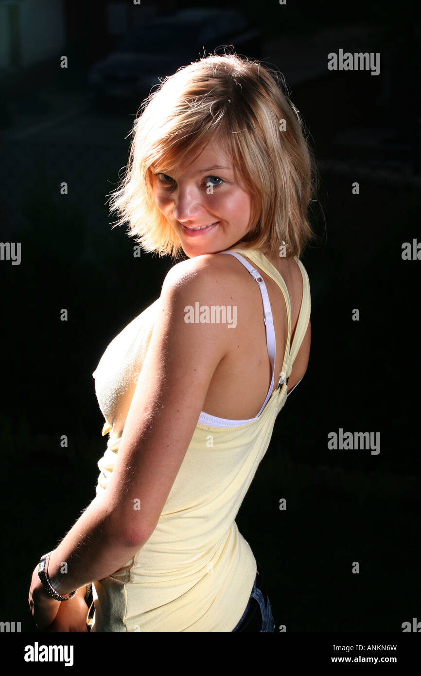 Blonde Frau in einem gelben Tanktop mit einem verspielten Lächeln, posiert draußen in sanftem Licht, ihr Haar fängt das Licht wunderschön ein. Stockfoto
