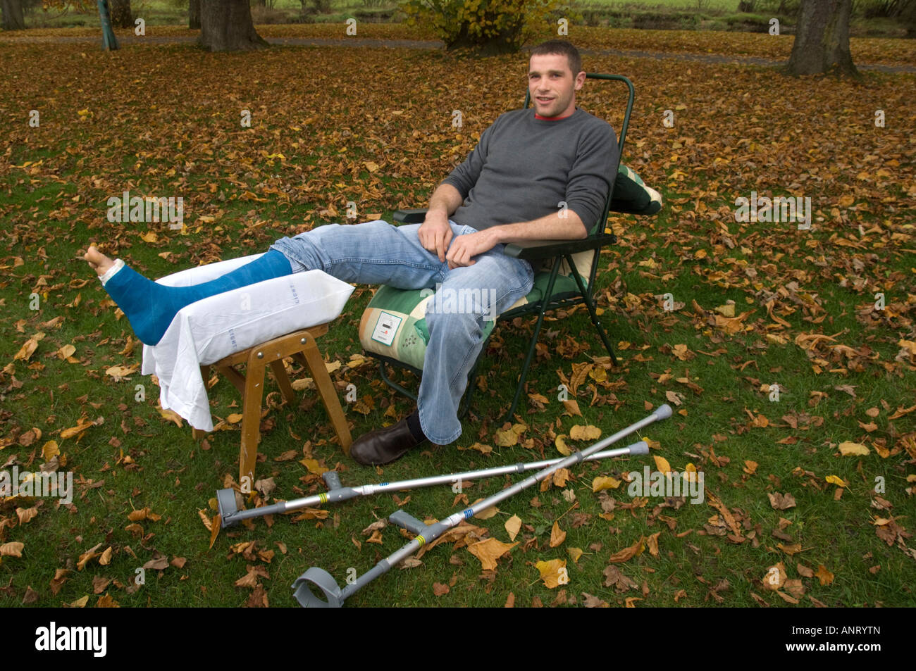 Junger Mann mit einem gebrochenen Bein in Gips, die im Freien auf einem Herbstnachmittag mit abgefallenen Blättern um ihn herum sitzen Stockfoto