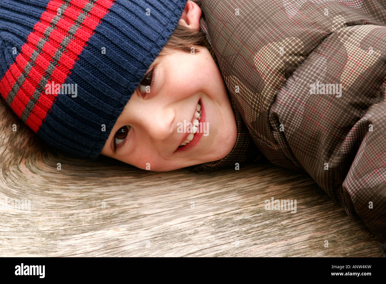 kleiner Junge mit Winter Hut und Mantel, auf einem Baumstamm liegend und lächelnd Stockfoto