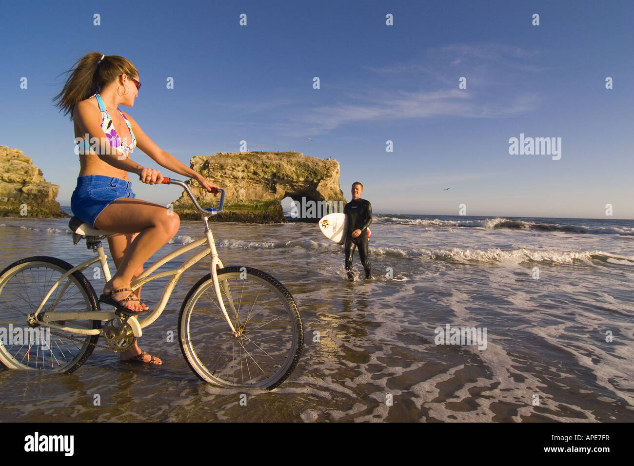 Eine Frau auf einem Cruiser-Fahrrad und ein Mann mit einem Surfbrett am Strand im Natural Bridges State Park in Santa Cruz in Kalifornien Stockfoto