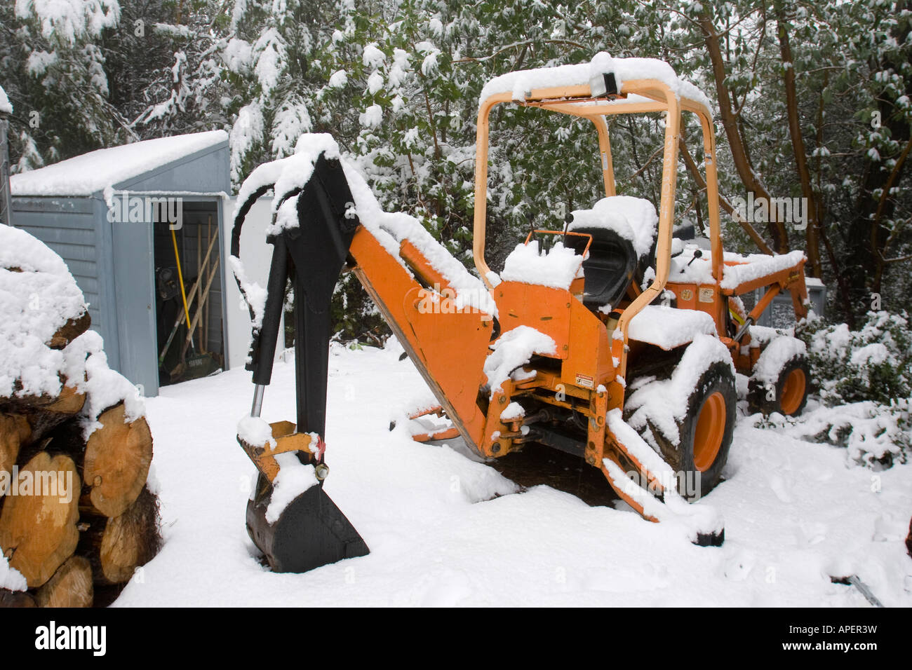 Schnee auf eine orange Traktor in einem ländlichen Winterszene mit Geräteschuppen und Stapel Brennholz gestapelt Stockfoto