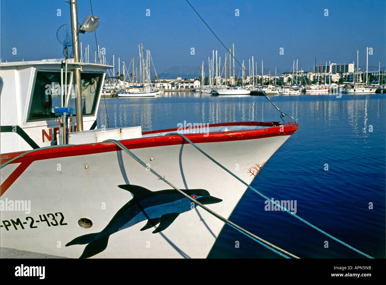 Ein Fischerboot sporting ein Bild eines springenden Delphins auf seine Bögen liegt in Ruhe mit der Marina und Fischerhafen von Port d Alcudia im Hintergrund Stockfoto