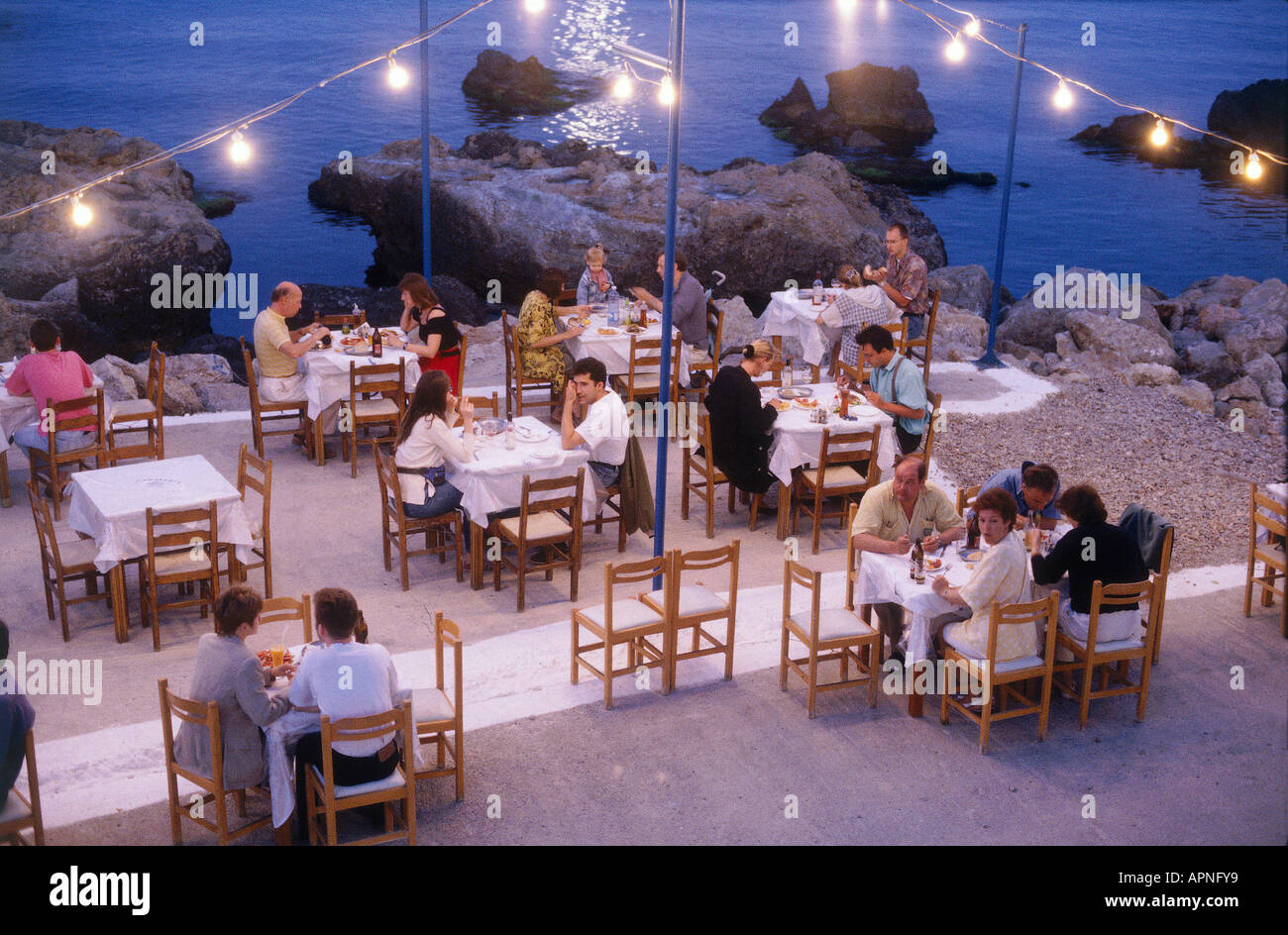 Menschen, die an den Tischen beim Abendessen in einer Wasser-Taverne, beleuchtet von Lichterketten im Hafen Stockfoto