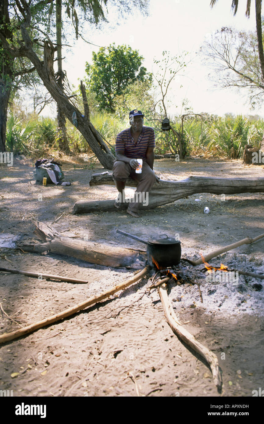 Lokale Mann sitzt auf einem Baumstumpf mit einem traditionellen schwarzen Kochtopf auf einem Feuer vor ihm. Stockfoto