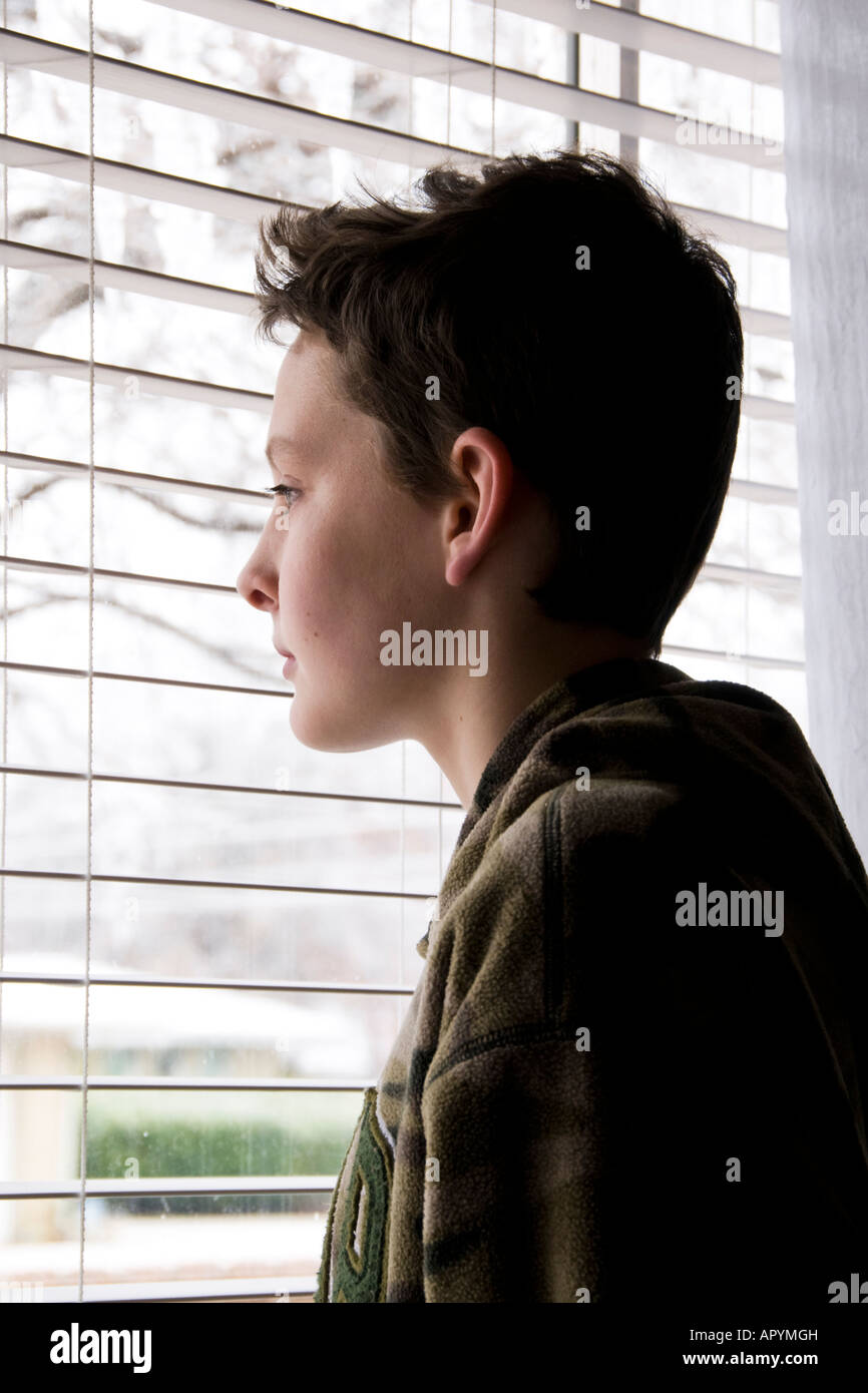 Ein 13-jähriger kaukasischer Junge, der traurig und einsam erscheint, steht vor einem Fenster mit Blick auf die Stadt. USA. Stockfoto