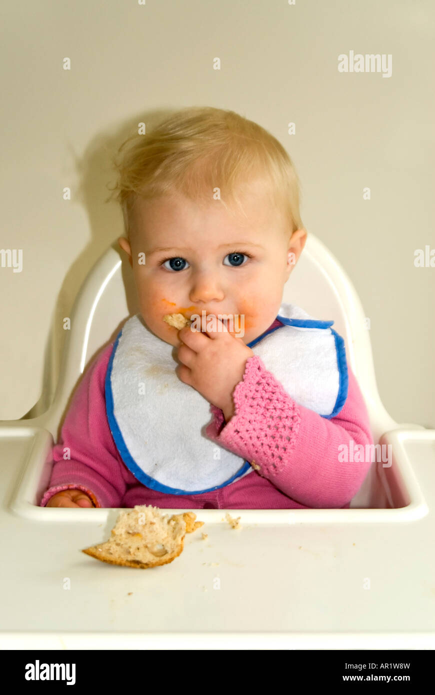 Vertikale Nahaufnahme von einem hübschen kaukasischen Babymädchen sitzt an ihrem Hochstuhl am Abend Fütterung selbst ein Stück Brot Stockfoto