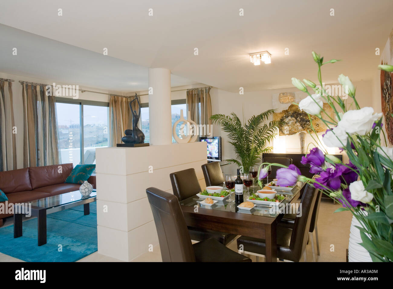 Innere des modernes Apartment mit Essbereich mit Salat, Suppe und Wein am Tisch mit Blumen im Vordergrund Stockfoto