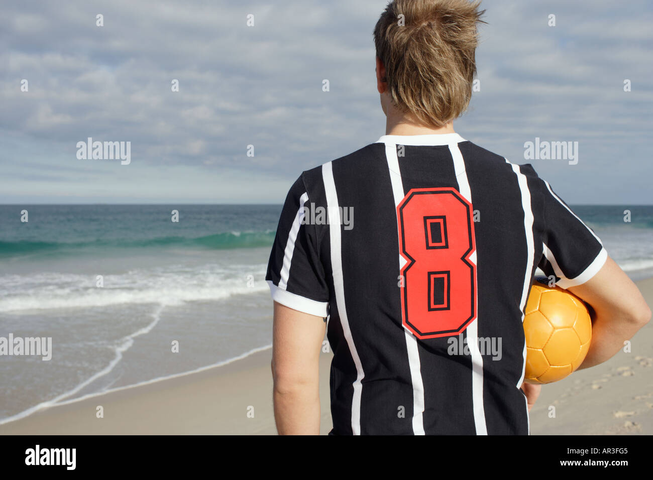 Junger Mann tragen Fußballtrikot halten Fußball Blick auf das Meer Stockfoto