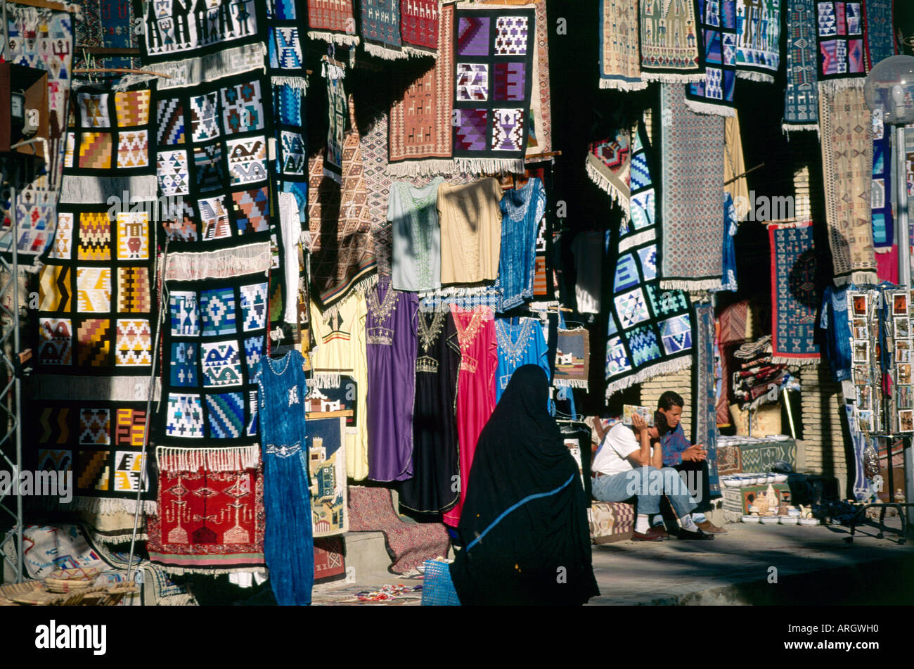 Eine lokale Frau gekleidet von Kopf bis Fuß in Schwarz geht ein paar Anbieter, die sitzen im Chat am Fuße eines Aufstands der bunte Teppiche und Kleider, die über die Backsteinfassaden der Geschäfte in der Avenue Habib Bourguiba Kaskade Stockfoto