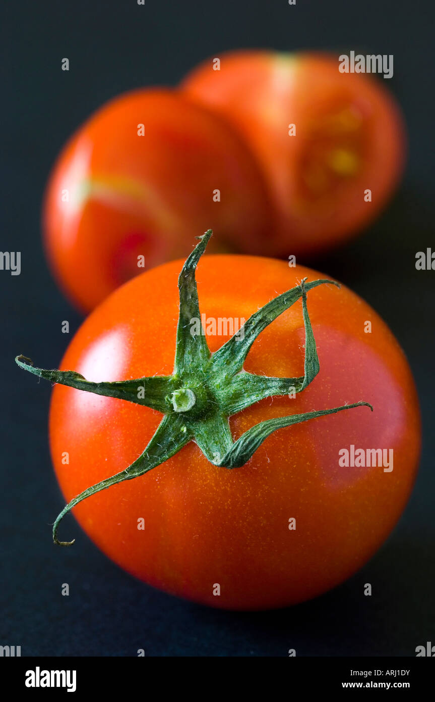 Ein Shinny rot unscharf gestellt Bio Tomate mit seinen Stengel auf gegen Schwarz mit geschnittenen Früchten im Hintergrund Stockfoto