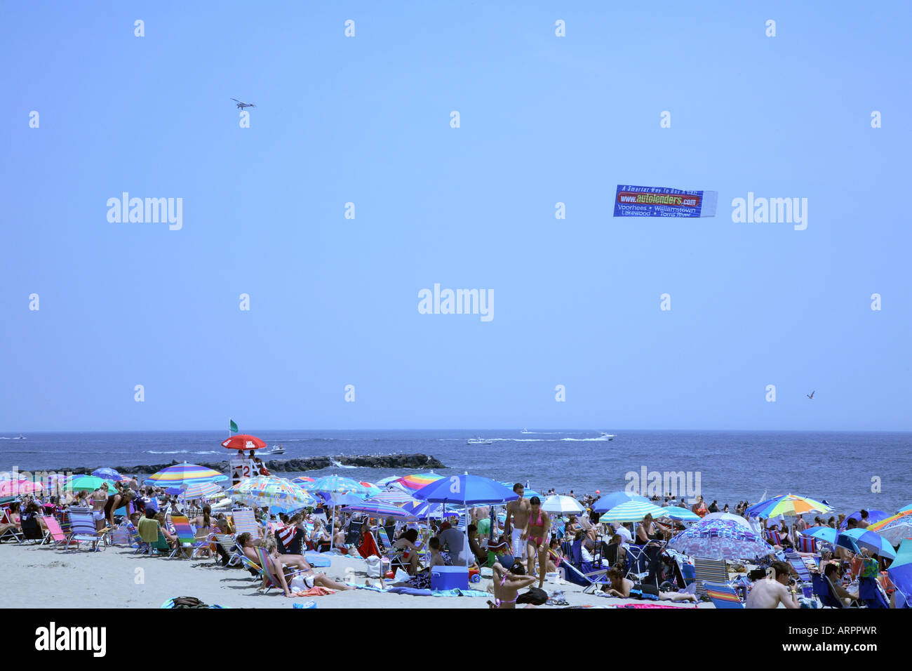 Masse von Menschen inmitten von großen bunten Sonnenschirmen mit Kleinflugzeug ziehen Werbebanner parallel zur Küste über Meer. Stockfoto