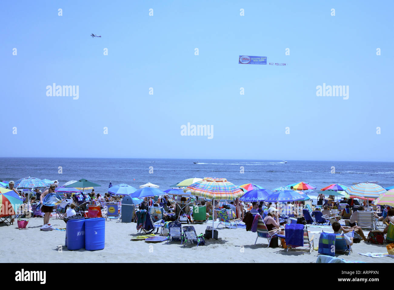 Masse von Menschen inmitten von großen bunten Sonnenschirmen mit Kleinflugzeug ziehen Werbebanner parallel zur Küste über Meer. Stockfoto