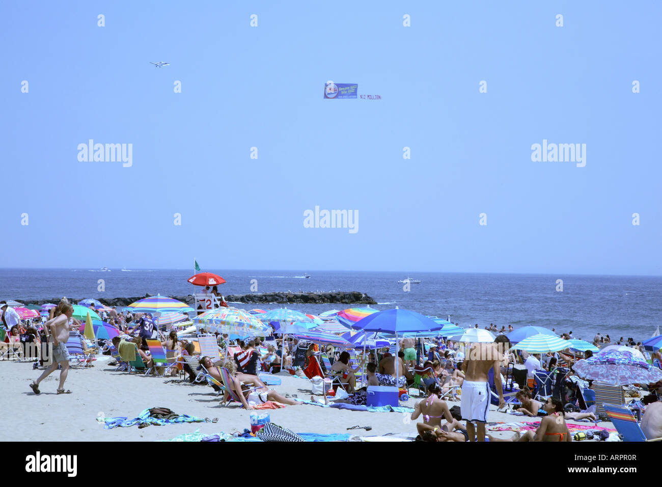 Masse von Menschen inmitten von großen bunten Sonnenschirmen mit Kleinflugzeug ziehen Werbebanner parallel zur Küste über Meer. Stockfoto