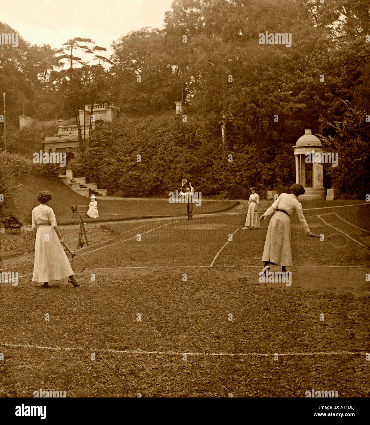 Ein vornehmendes Tennisspiel in Großbritannien um 1900. Ein Mann und 3 Damen spielen in einem Landhaus/Herrenhaus auf einem unebenen Rasen – ein Zeitvertreib der Oberschicht. Stockfoto