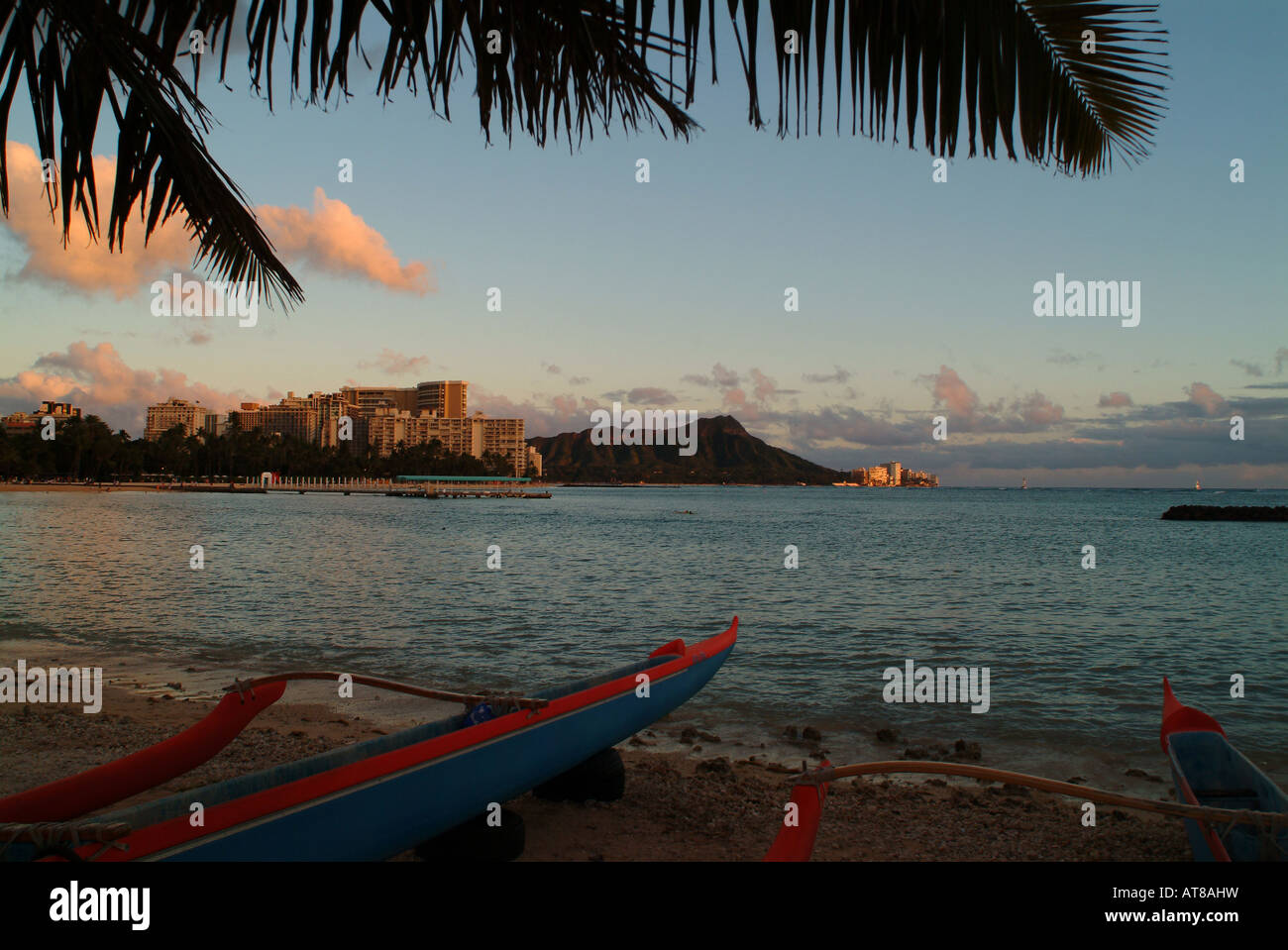 Der vordere Rumpf von blauen und roten Kanus säumen den Sand während des Sonnenuntergangs am Waikiki Beach mit Diamond Head in der Ferne. Stockfoto