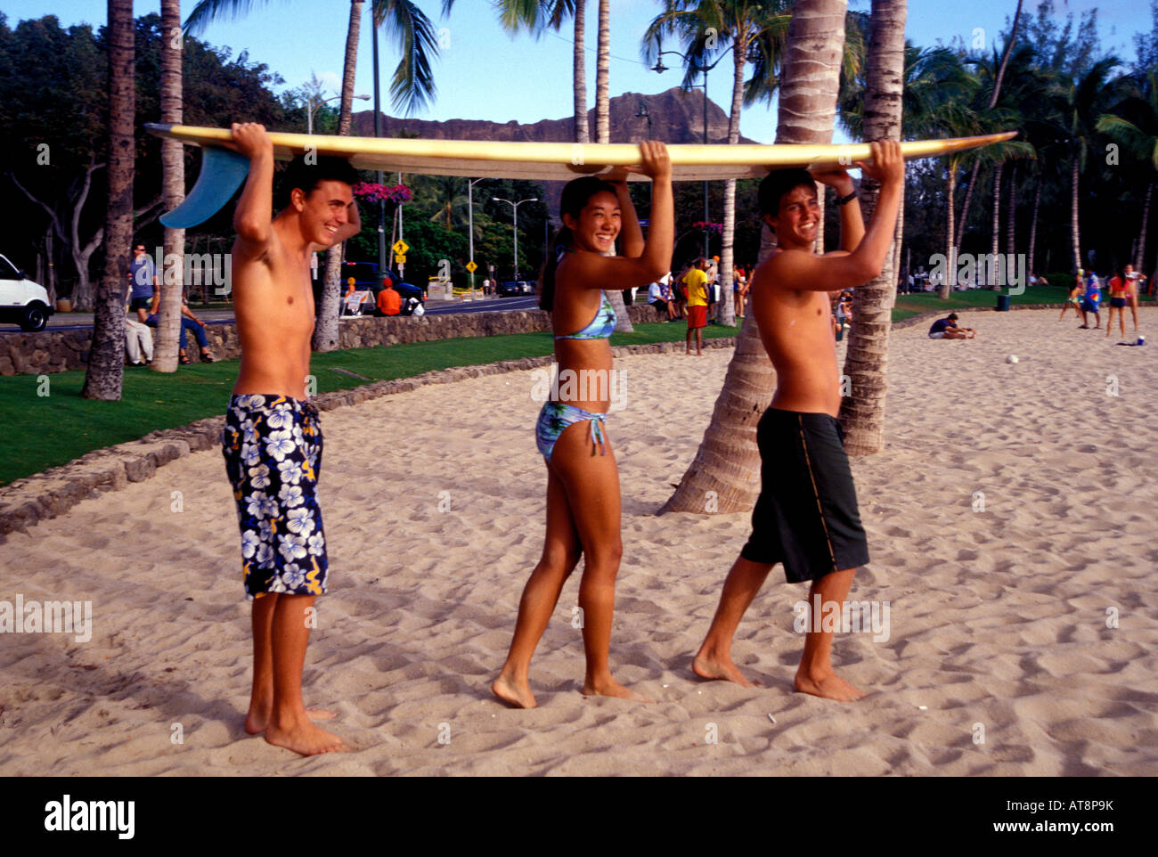 Drei Jugendliche Lächeln, als sie ein Surfbrett auf dem Kopf in den Sand am Strand von Waikiki balancieren. Stockfoto