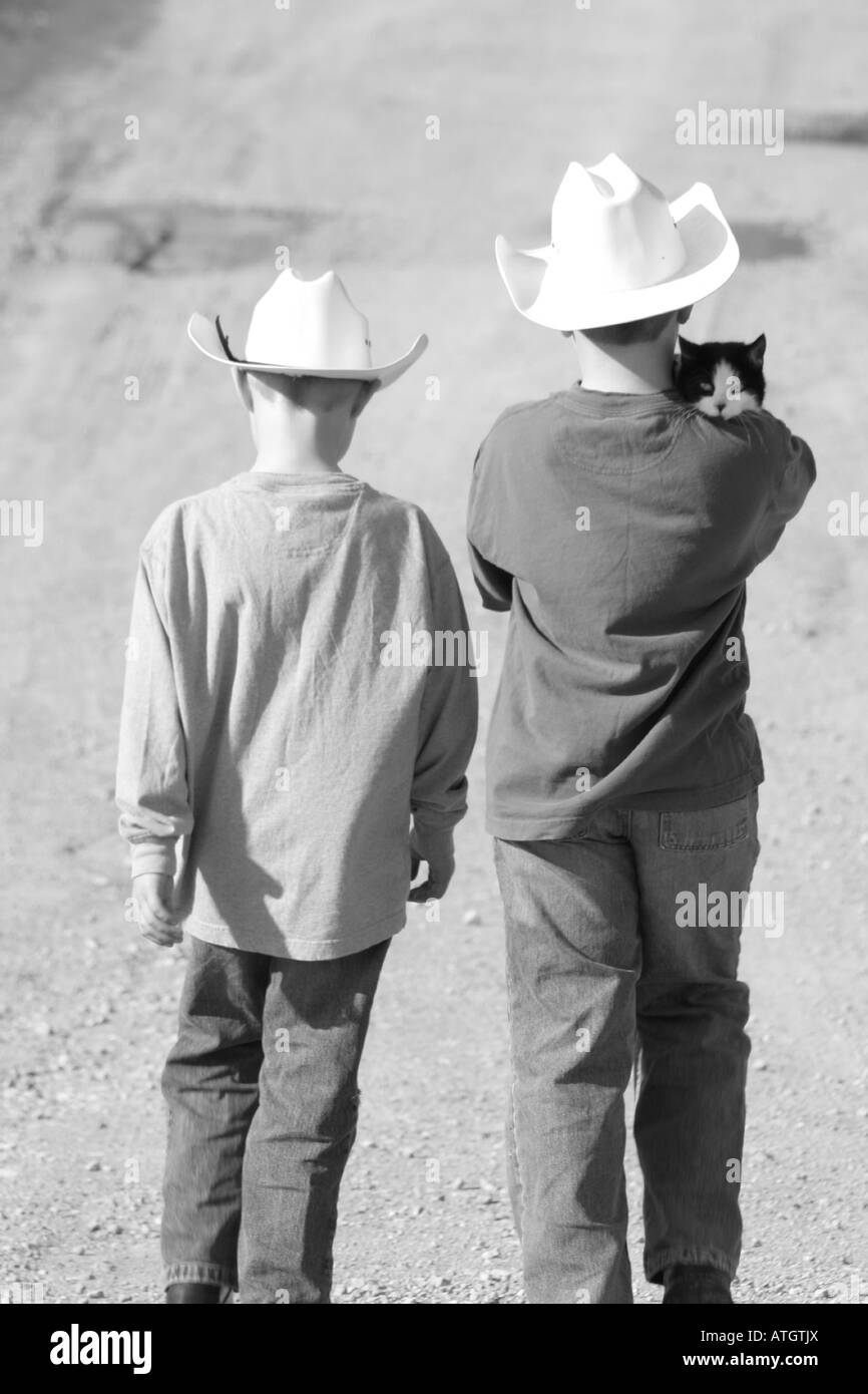 zwei jungen mit Cowboy-Hüte zu Fuß Gasse ein Bauernhof mit Katze auf jungen Schultern Stockfoto