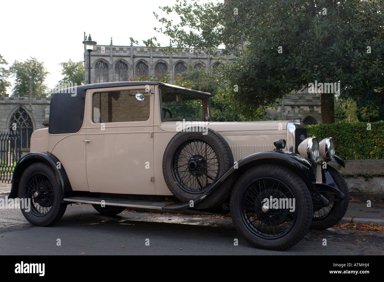 Oldtimer in Creme und Schwarz, der auf einer Straße in der Nähe einer historischen Steinkirche geparkt wurde, spiegelt die Automobilgeschichte und die Eleganz der Epoche Großbritanniens wider Stockfoto