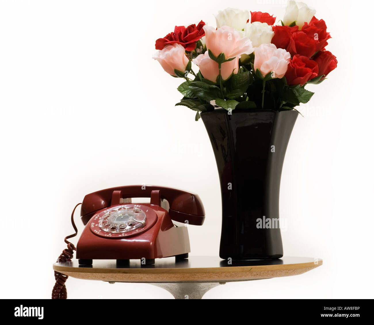 Kleiner Beistelltisch mit einer schwarzen Vase voller rote und rosa Rosen neben einem alten Stil Retro-rotary Telefon über weiß Stockfoto
