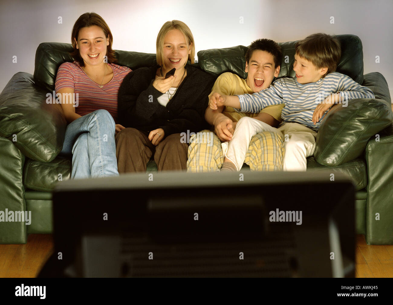 Drei Jugendliche und ein Kind sitzt auf grüne Couch zusammen, unscharf lachen, Rückansicht des tv im Vordergrund Stockfoto