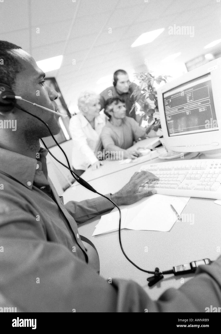 Mann mit Kopfhörer mit Computer, Kolleginnen und Kollegen in der Hintergrund jedoch unscharf, B&W Stockfoto
