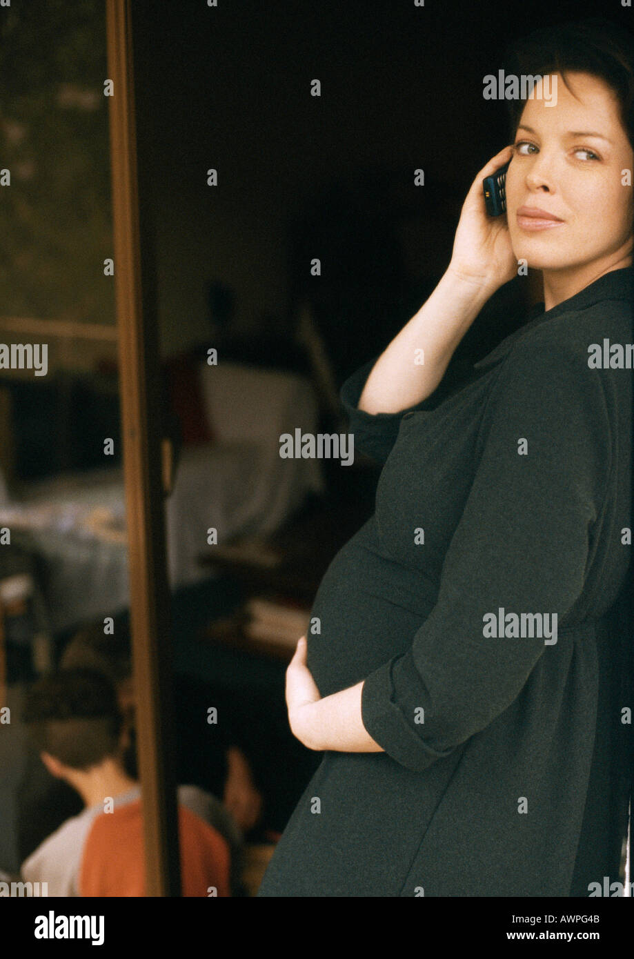 Schwangere Frau stehend, mit Handy, Kind sitzt am Boden im Hintergrund Stockfoto