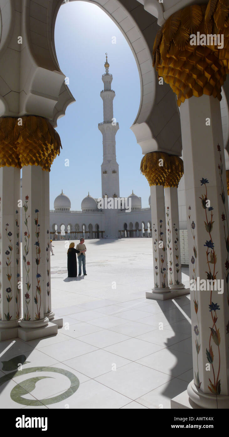Ein Minarett, das durch einen Torbogen gesehen wird, während ein Paar im riesigen 17.000 Quadratmeter großen Innenhof der Sheikh Zayed Moschee in Abu Dhabi, VAE steht. Stockfoto