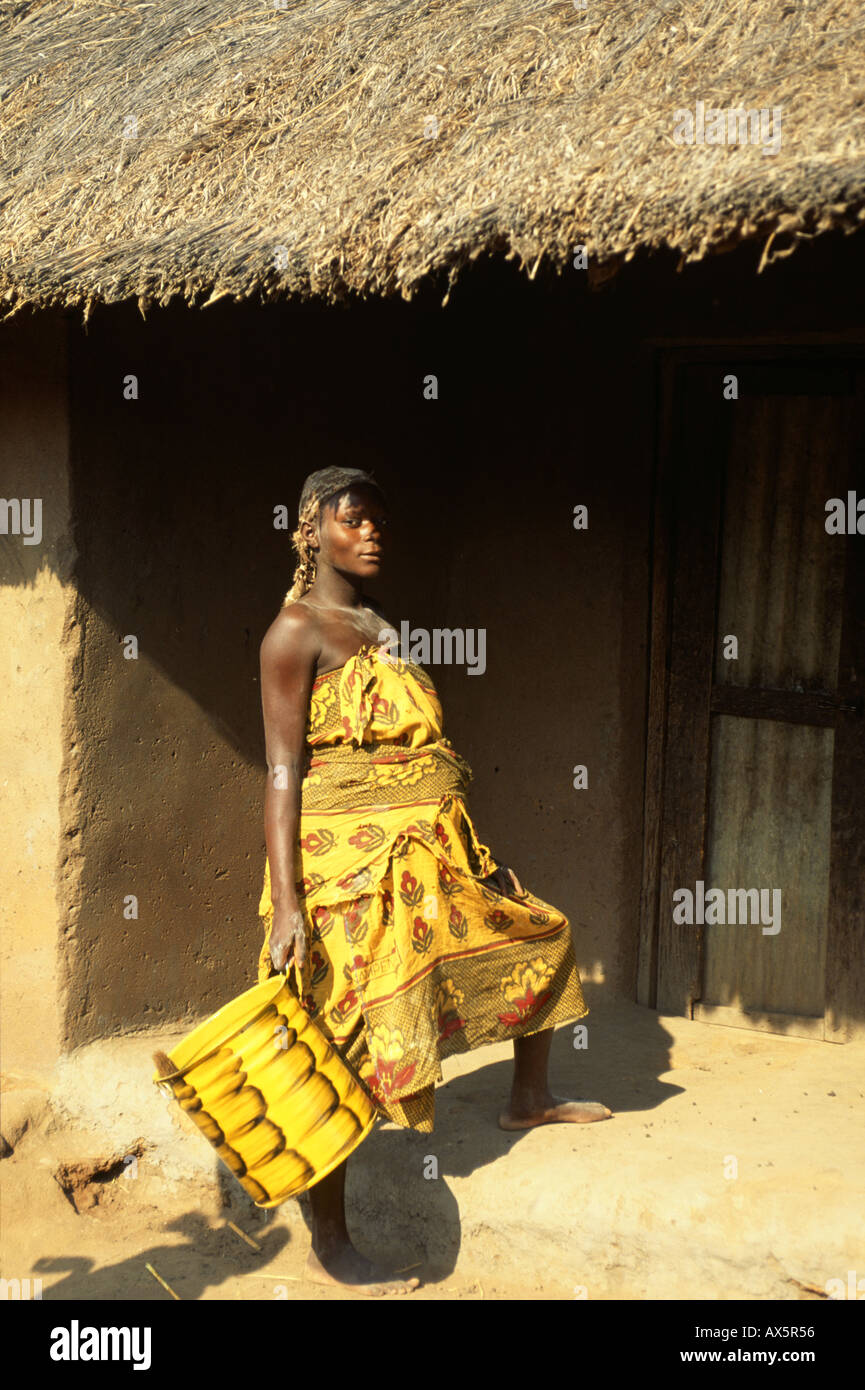 Yumba, Tansania. Frau zerlumpten bedruckter Baumwolle Kleidung tragen eine gelbe kann außerhalb einer Adobe und strohgedeckten Hütte. Stockfoto