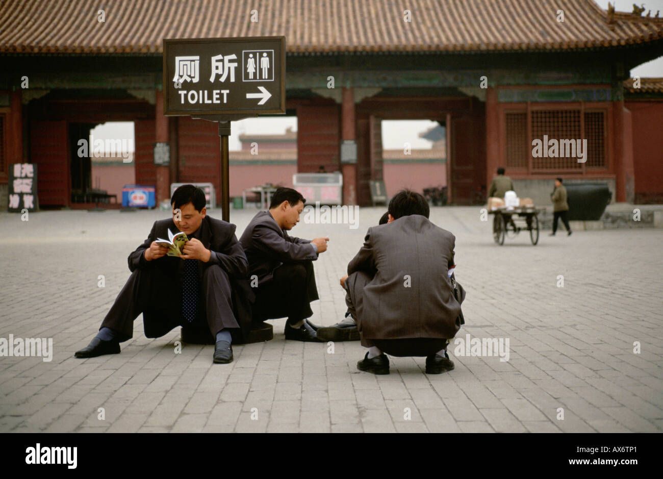 Chinesische Männer hocken und lesen Sie Bücher neben ein Zeichen in chinesischen Schriftzeichen, die unter Angabe einer Toilette wird Neary durch an die Verbotene Stadt Stockfoto