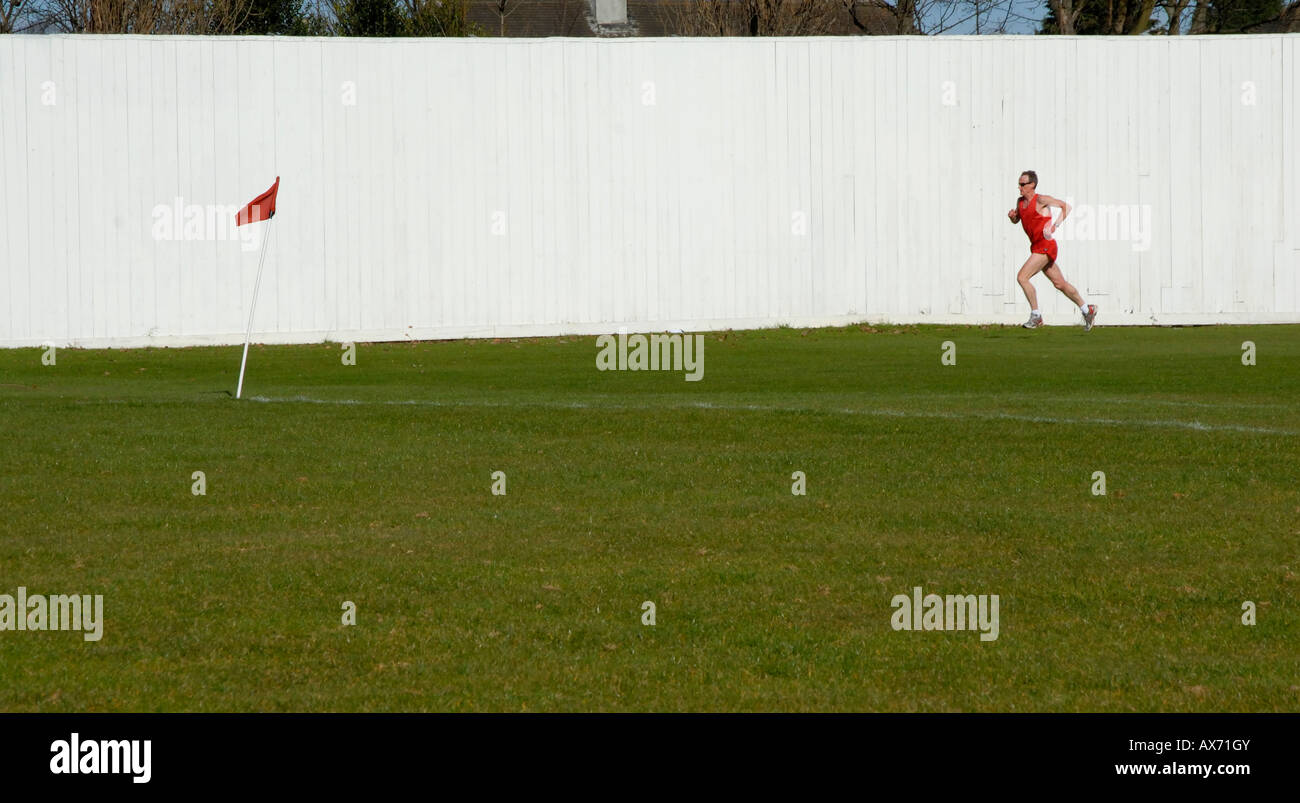 Mann in rote Shorts und Top Joggen auf ein Cricket-Club-Platz vorbei an weißen Zaun in Richtung rot Eckfahne, Cheam, Südlondon, Surrey Stockfoto